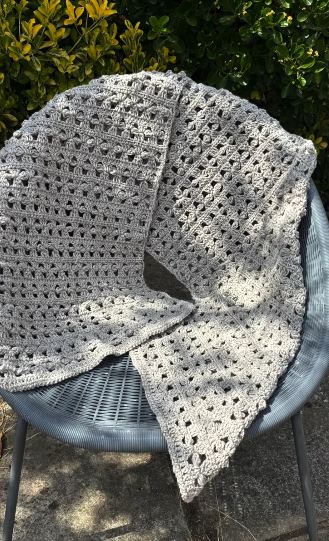 Cloverline Alignment Shawl, a long rectangular crochet shawl in natural silver-grey wool with rows of clover-shaped stitches, draped over a garden chair in sunlight