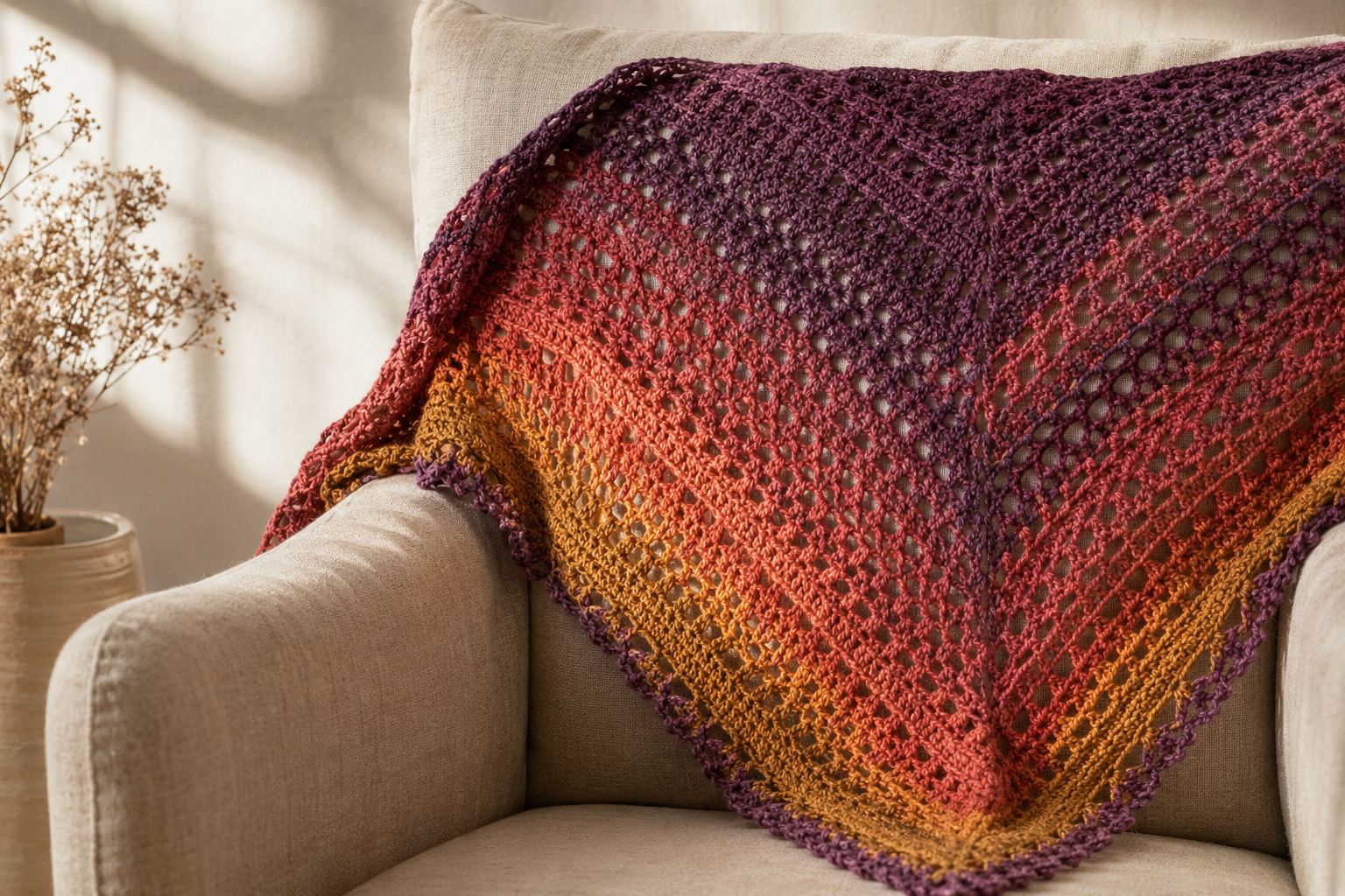 Plum Orchard Shawl, a triangular hand-crocheted shawl fading from deep plum through coral to saffron, draped over a linen armchair