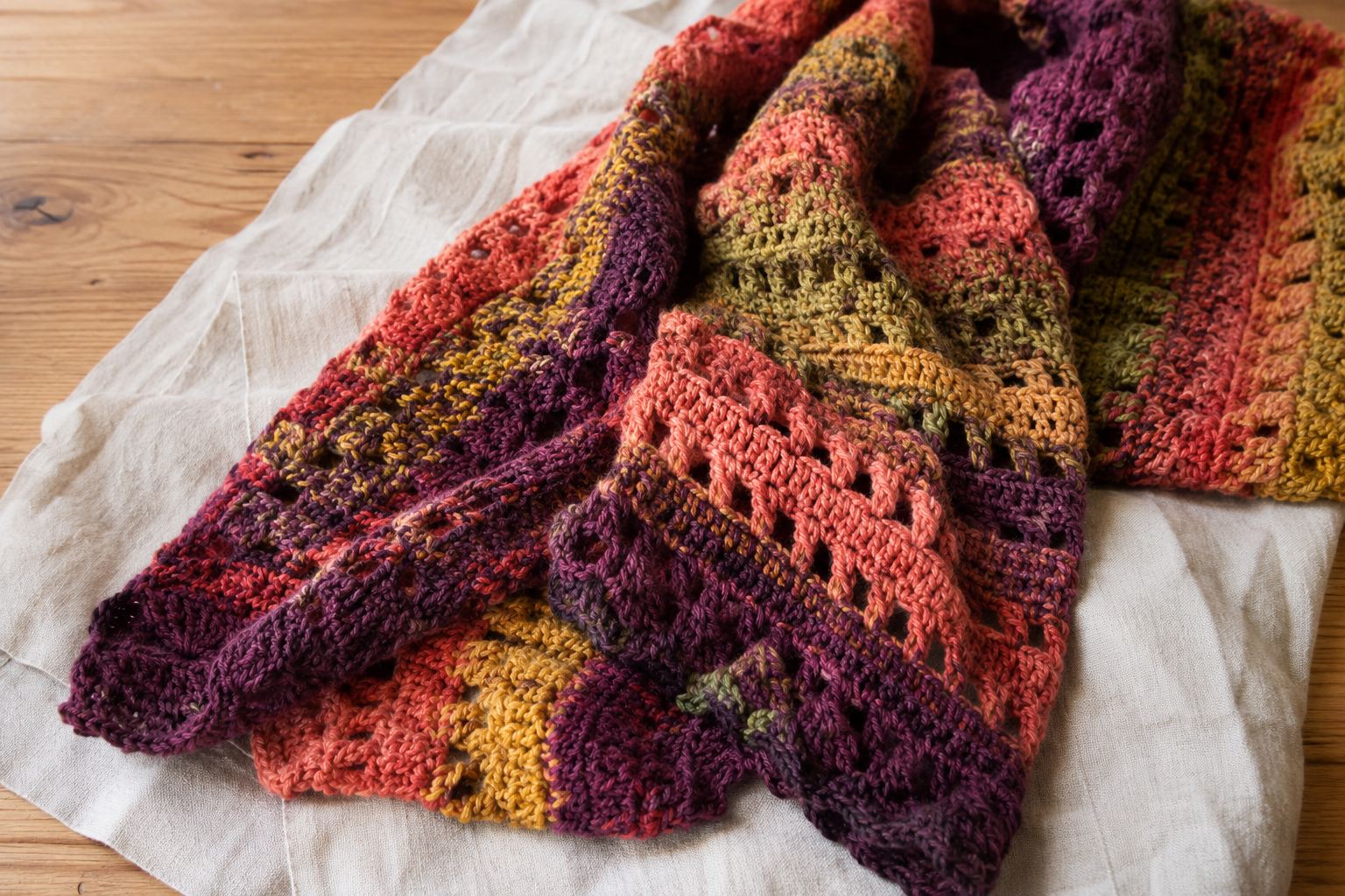 Sunlit Hearth Scarf, an oversized crochet scarf in stripes of saffron, moss green, coral and plum, folded on a linen cloth on a wooden table